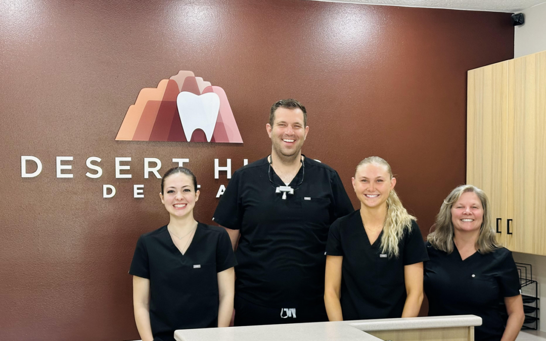 A team of four dental professionals posing for a photo at the reception of Desert Hills Dental. The team consists of two women and two men, wearing black dental scrubs, standing behind the reception counter with a logo featuring a mountain and tooth in the background.